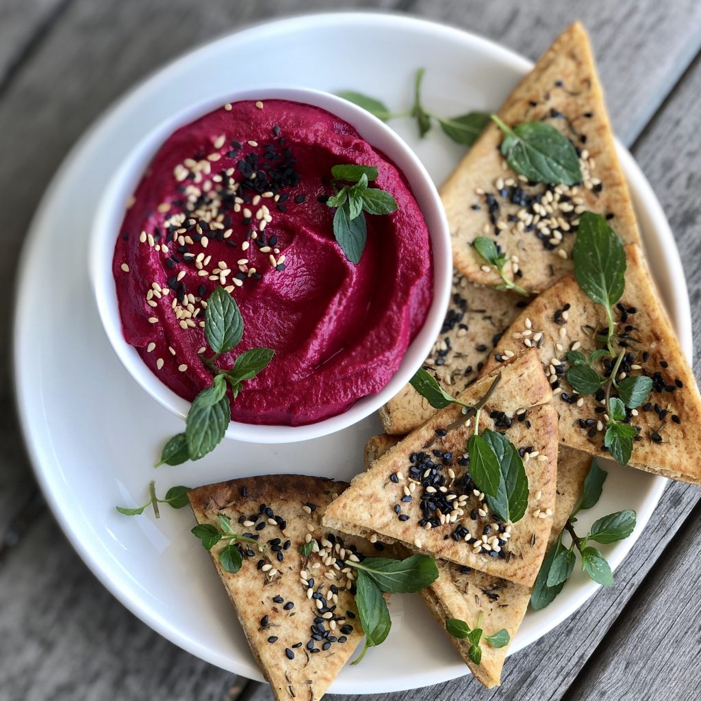 Roasted Beet Dip in a white bowl on a white plate with seeded pita chips to the side. 