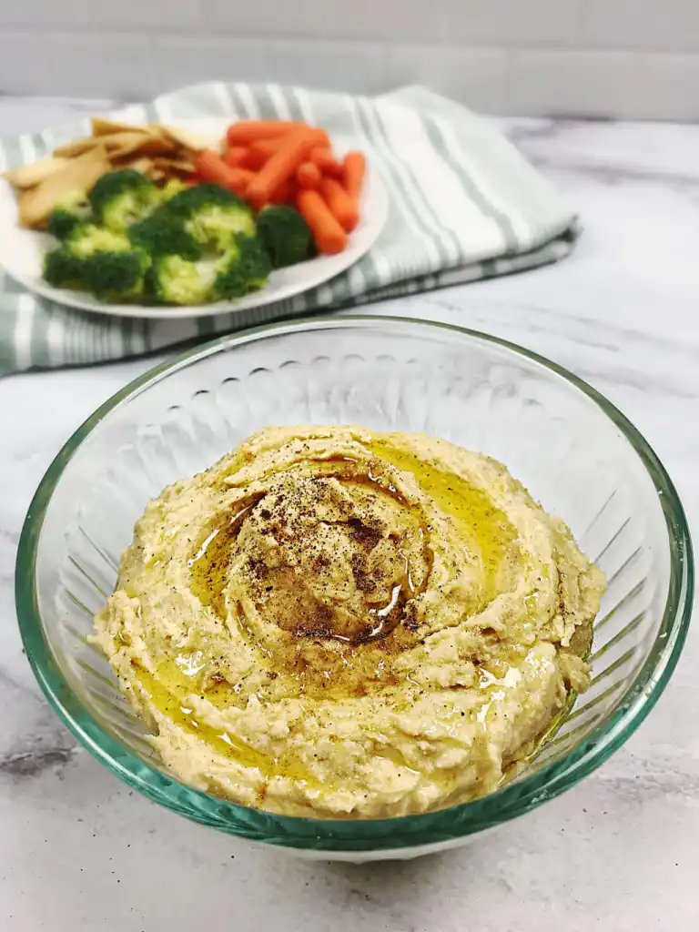 Flaxseed Hummus in a glass bowl to the right top corner of the image is a plate with veggies and pita chips. 