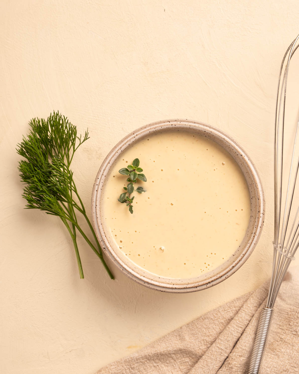 A stoneware bowl with a cream Horseradish Aioli along side some bright herbs and a whisk.