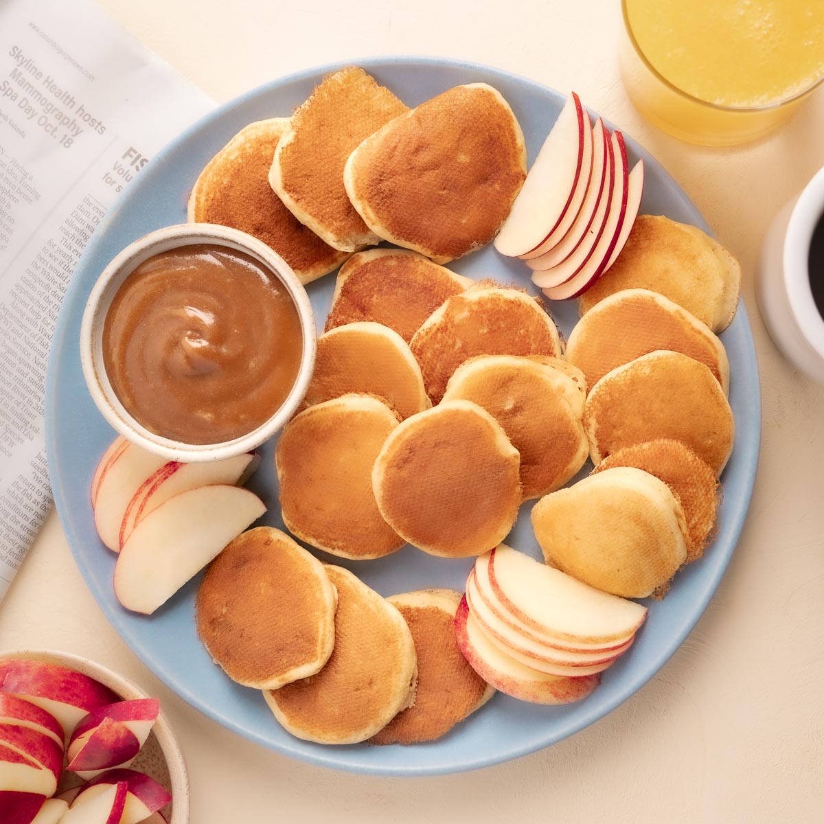 Mini Pancakes on a blue plate with Vanilla Cinnamon Peanut Butter Maple Syrup in a small white bowl on the plate. Red sliced apples are also shown as dippers for the peanut butter syrup. 