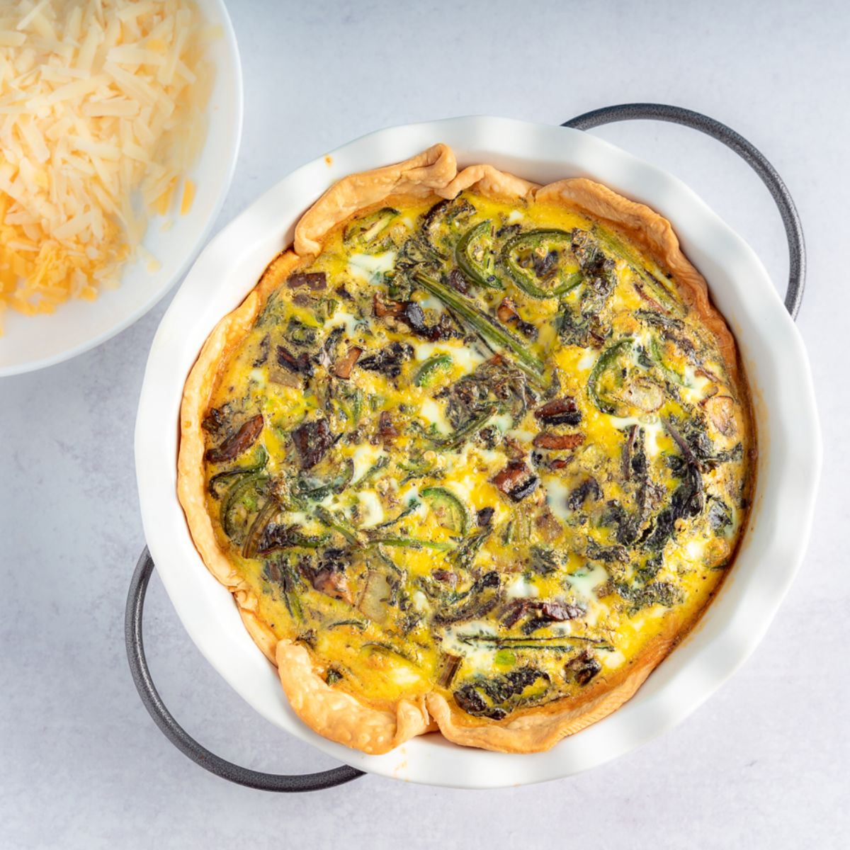 Mushroom Quiche in a white pie pan with shredded cheeses to the left side of the image
