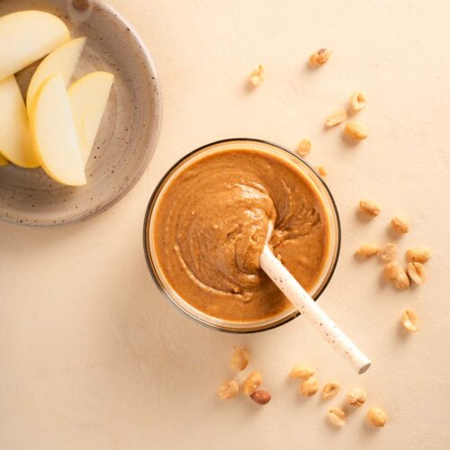 Nut butter in a glass bowl with a ceramix spoon and some peanuts scattered around the ground, sliced golden apples i the top left corner.