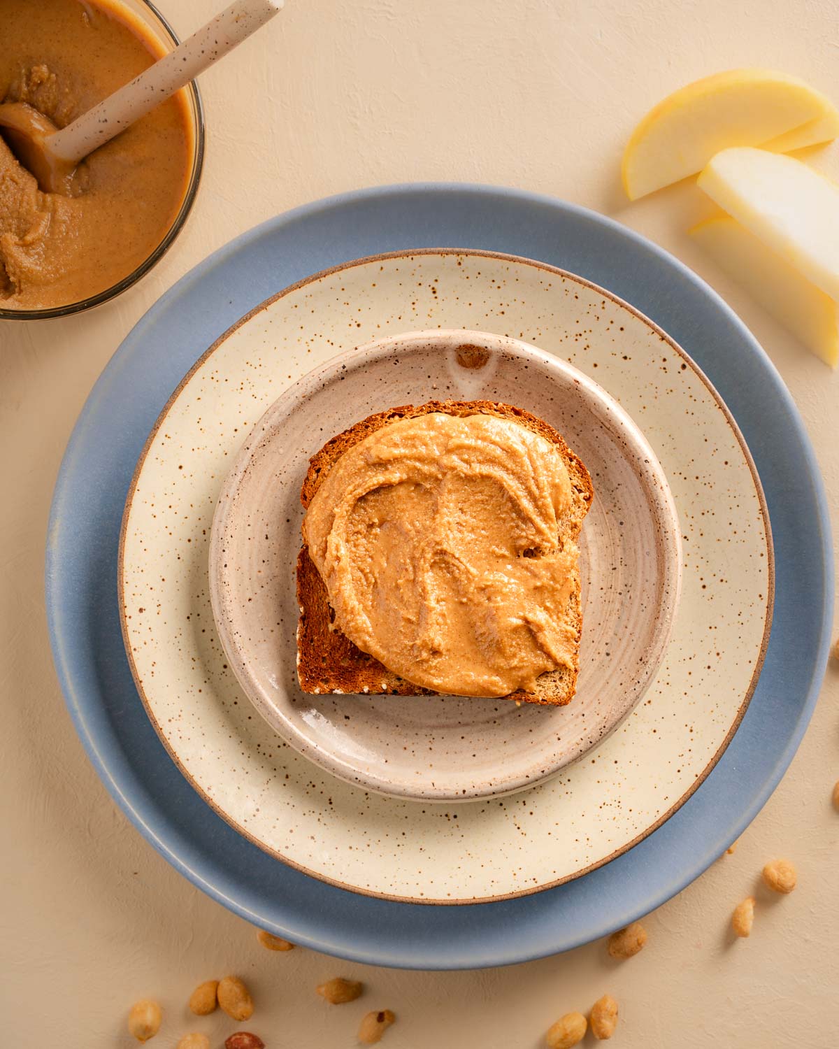 A slice of bread lathered with Vanilla Cinnamon Peanut Butter sitting on top of three stacked plates of different sizes. Food props include the bowl of nut butter with a ceramic spoon, sliced apples and peanuts. 