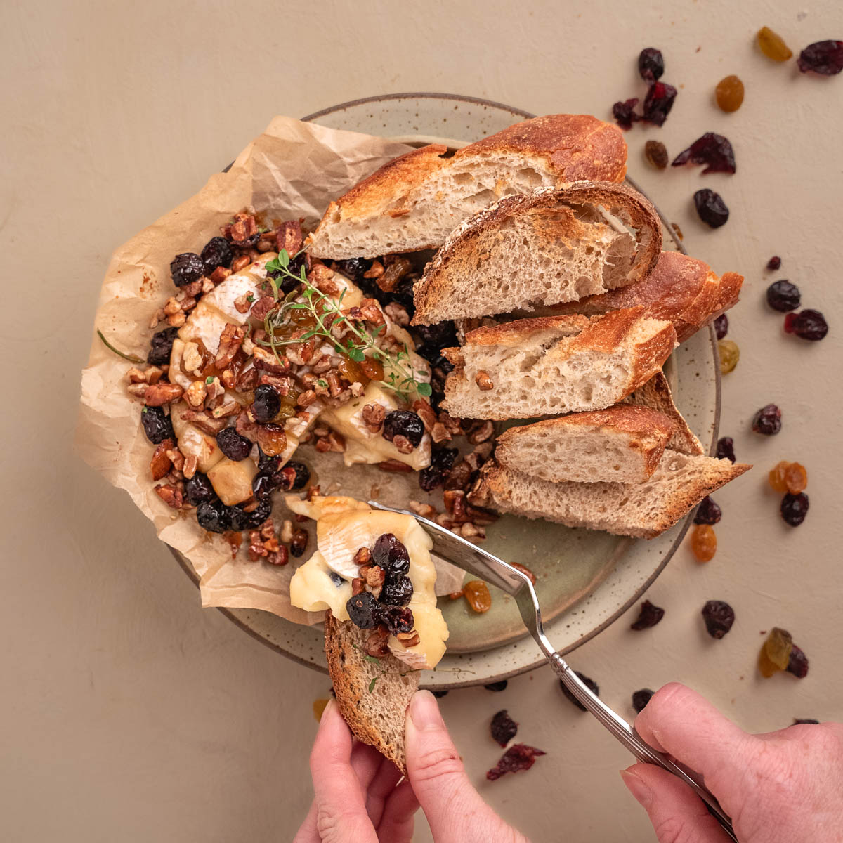 Camembert Cheese on stacked plates with sliced bread topped with nuts and dried fruit. A pair of hands is slicing into the cheese and spreading it along bread. 