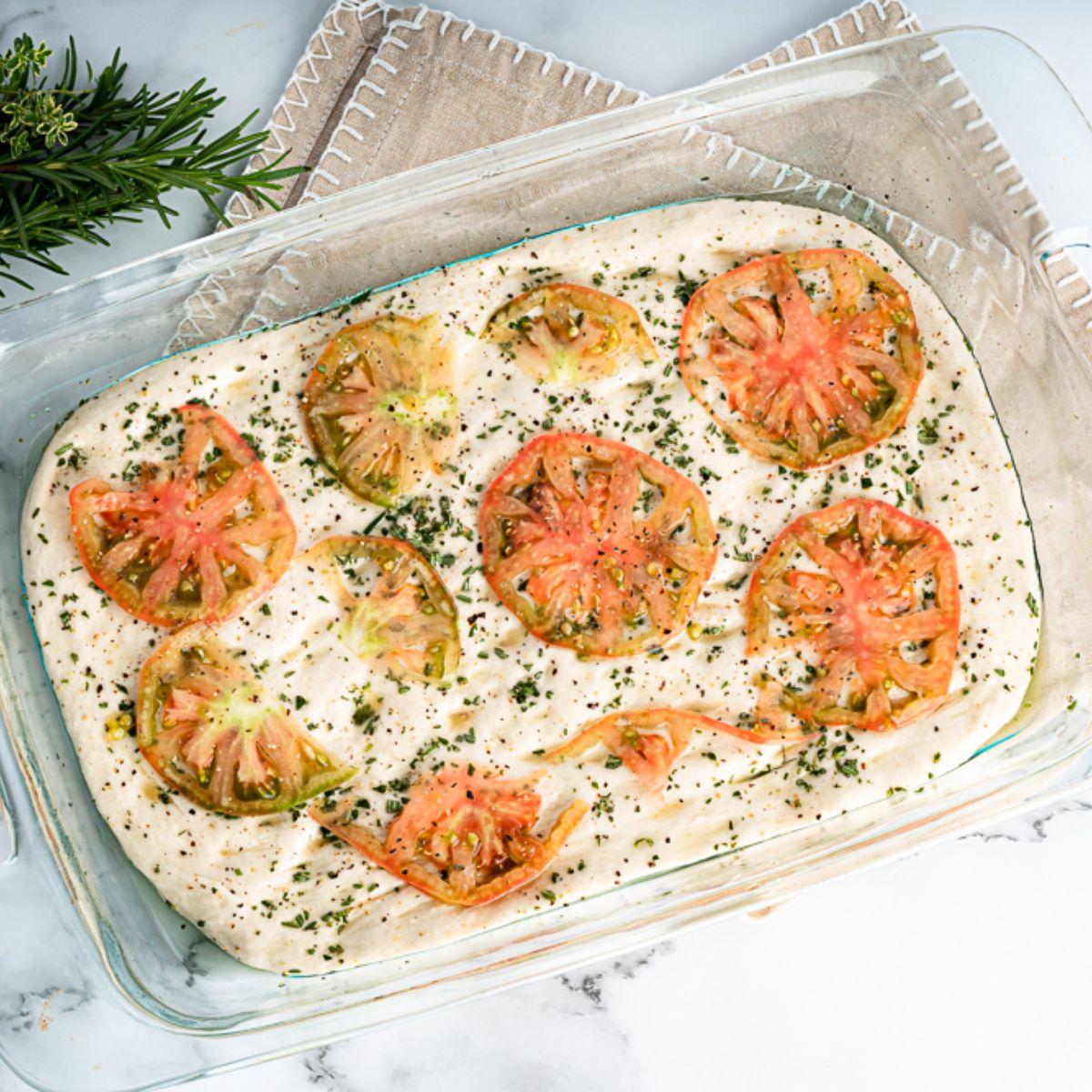 Uncooked focaccia in a glass pan with sliced tomatoes and herbs to the left top corner
