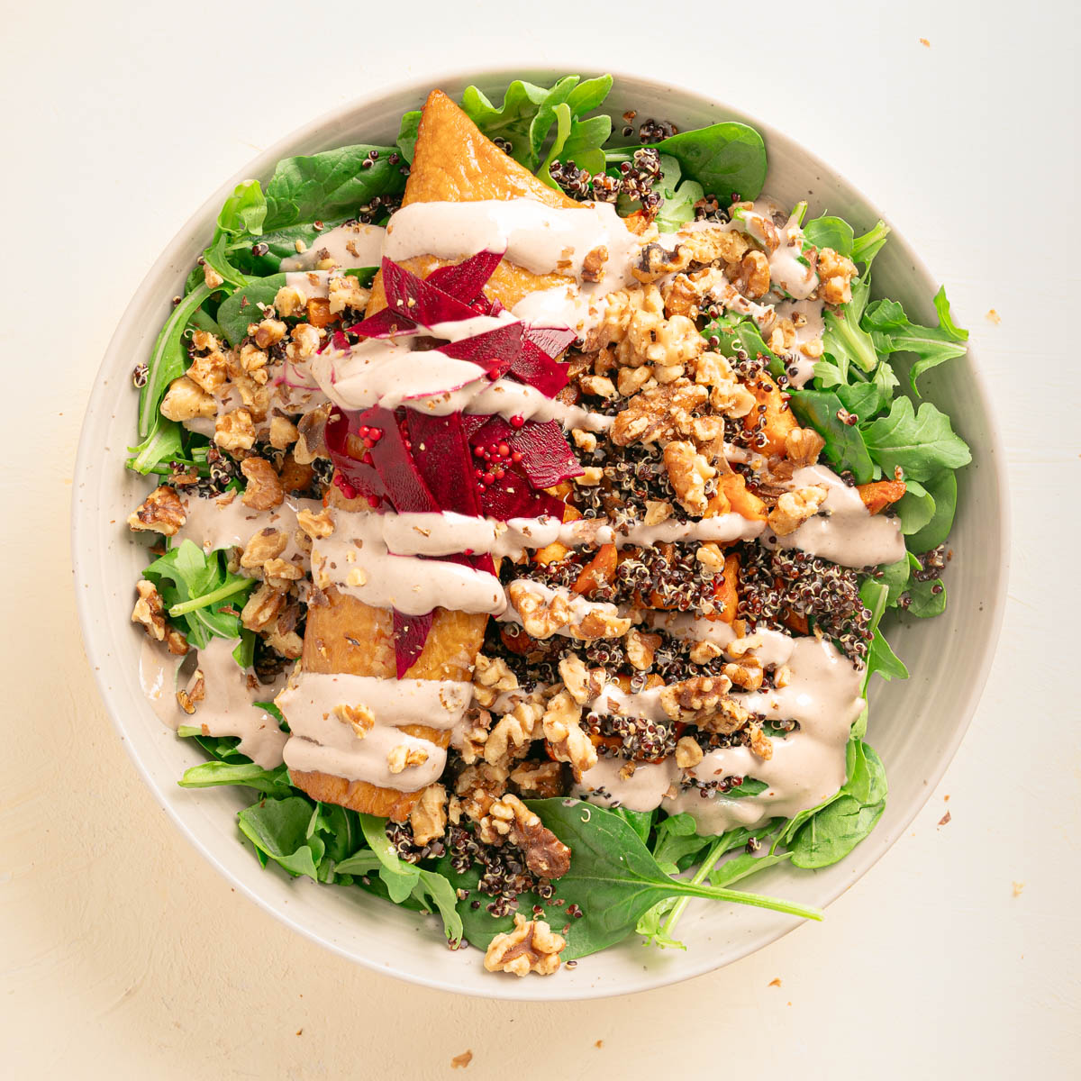 A stoneware bowl with arugula and spinach salad topped with toasted vegetables, quinoa, smoked fish, pickled beets, toasted walnuts, and a creamy walnut dressing.