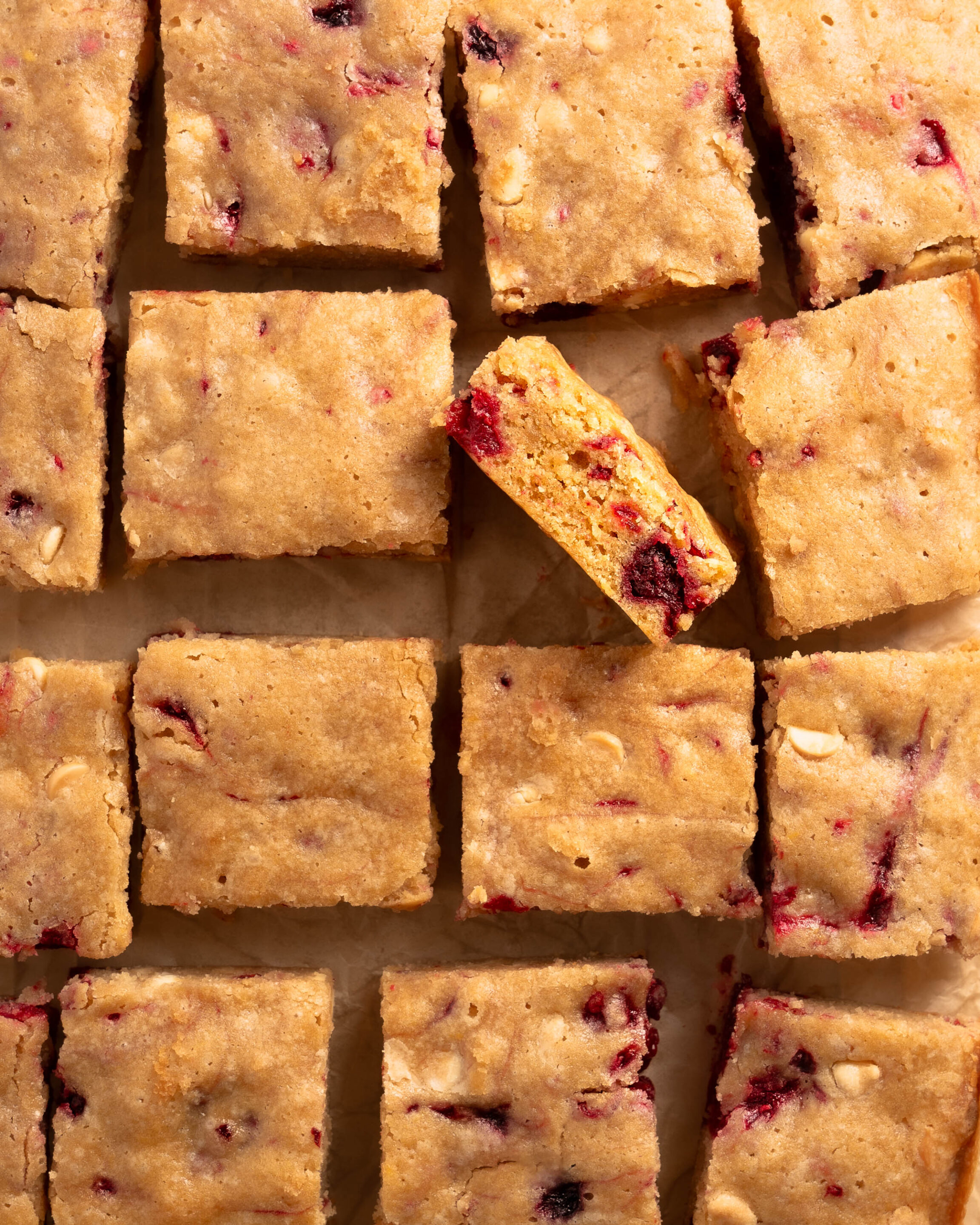 Raspberry White Chocolate Blondies on a wire wrack covered in light brown parchment paper. One blondie is turned on its side to show the chunks of raspberry and white chocolate.