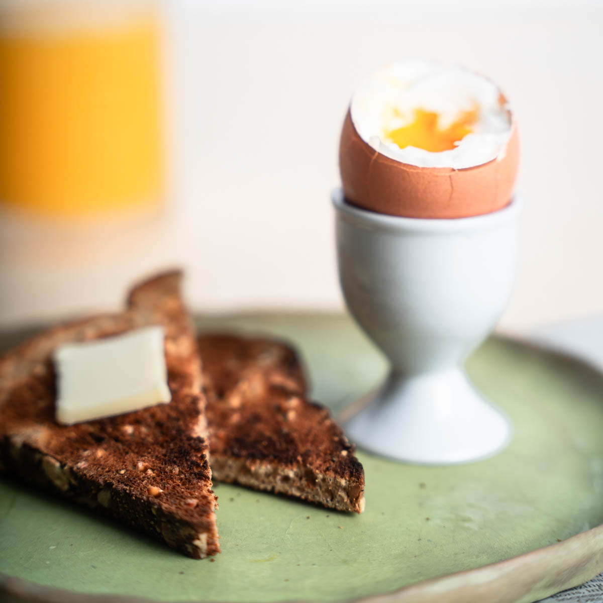 Soft cooked egg in a egg holder with toast on a sage green plate, and orange juice in the background.