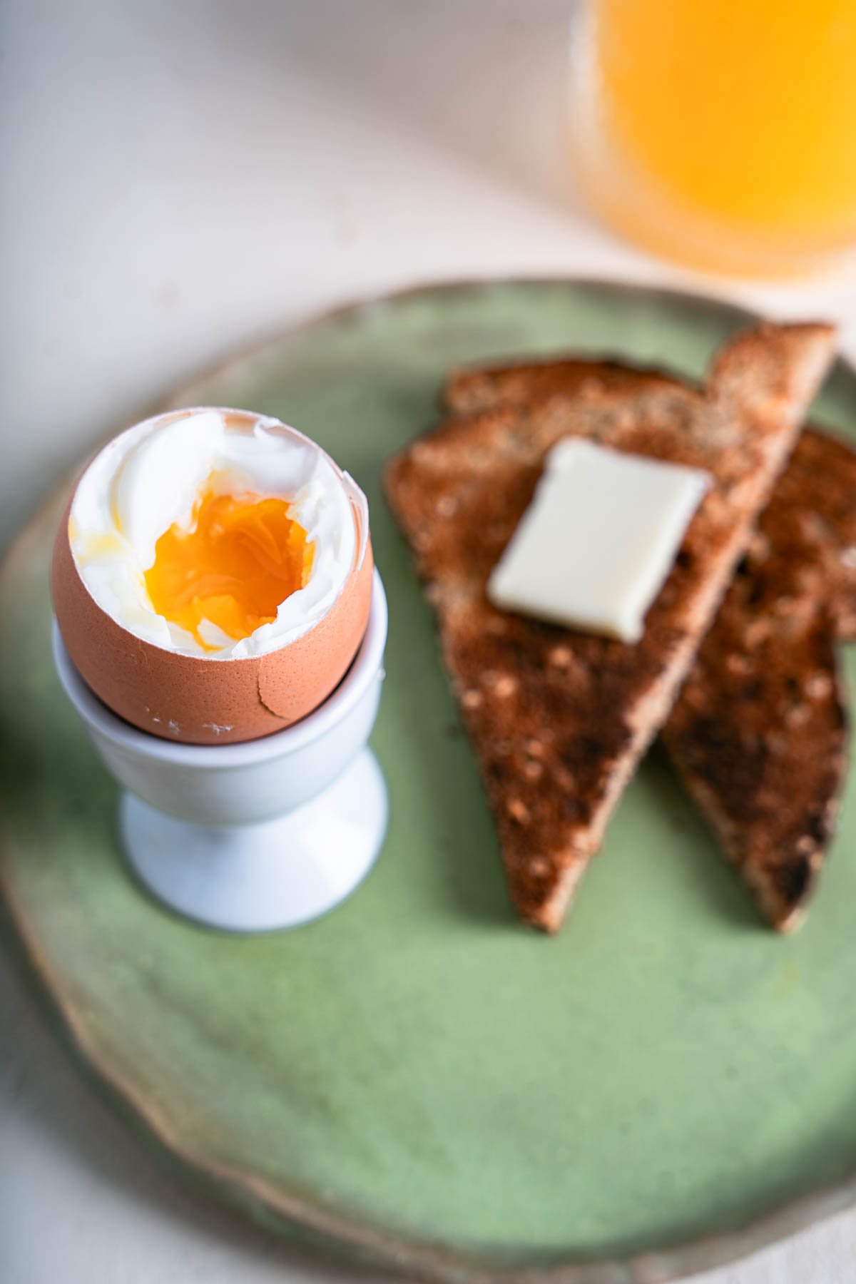 Soft Cooked Egg in a white egg holder with toast and butter on a sage green plate.