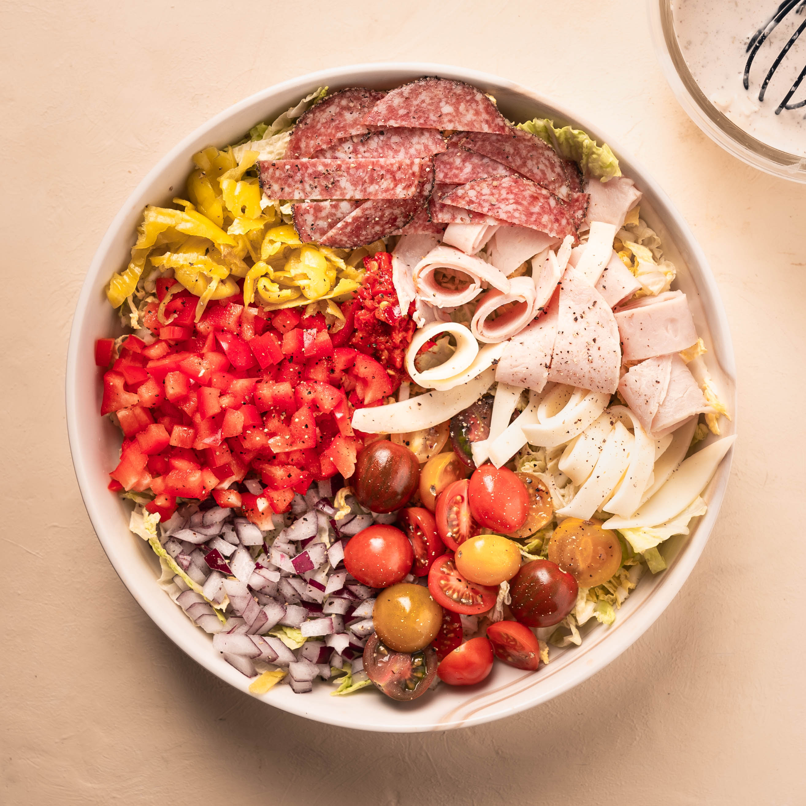 A Grinder Salad in a white bowl with deli meats, provolone cheese, various peppers, red onion, and tomatoes.