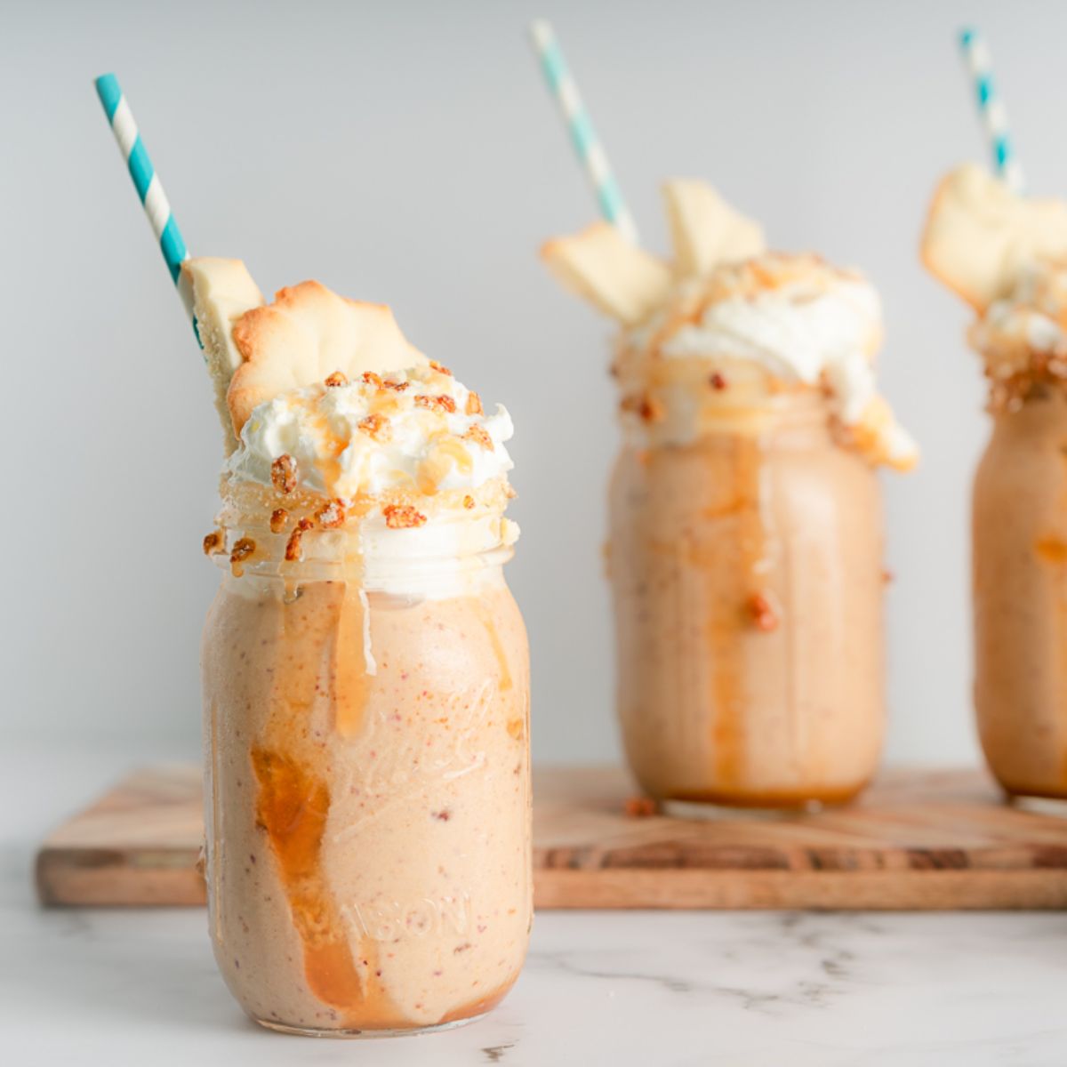Three peach cobbler protein shakes with one in front and the other two raised on a wooden serving board behind. All garnished with whipped cream, shortbread cookies, caramel, and a straw. 