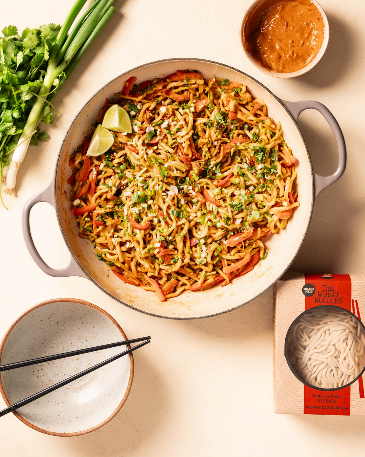 Trader Joes Thai Wheat Noodle PEanut Stir Fry in a pan with the package of noodles, a bowl with chopsticks, herbs, and peanut sauce as props surrounding the dish.