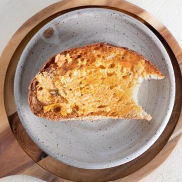 Tomato herb butter smeared on a piece of crust sourdough bread placed on a speckled stoneware plate on a circular brown serving platter