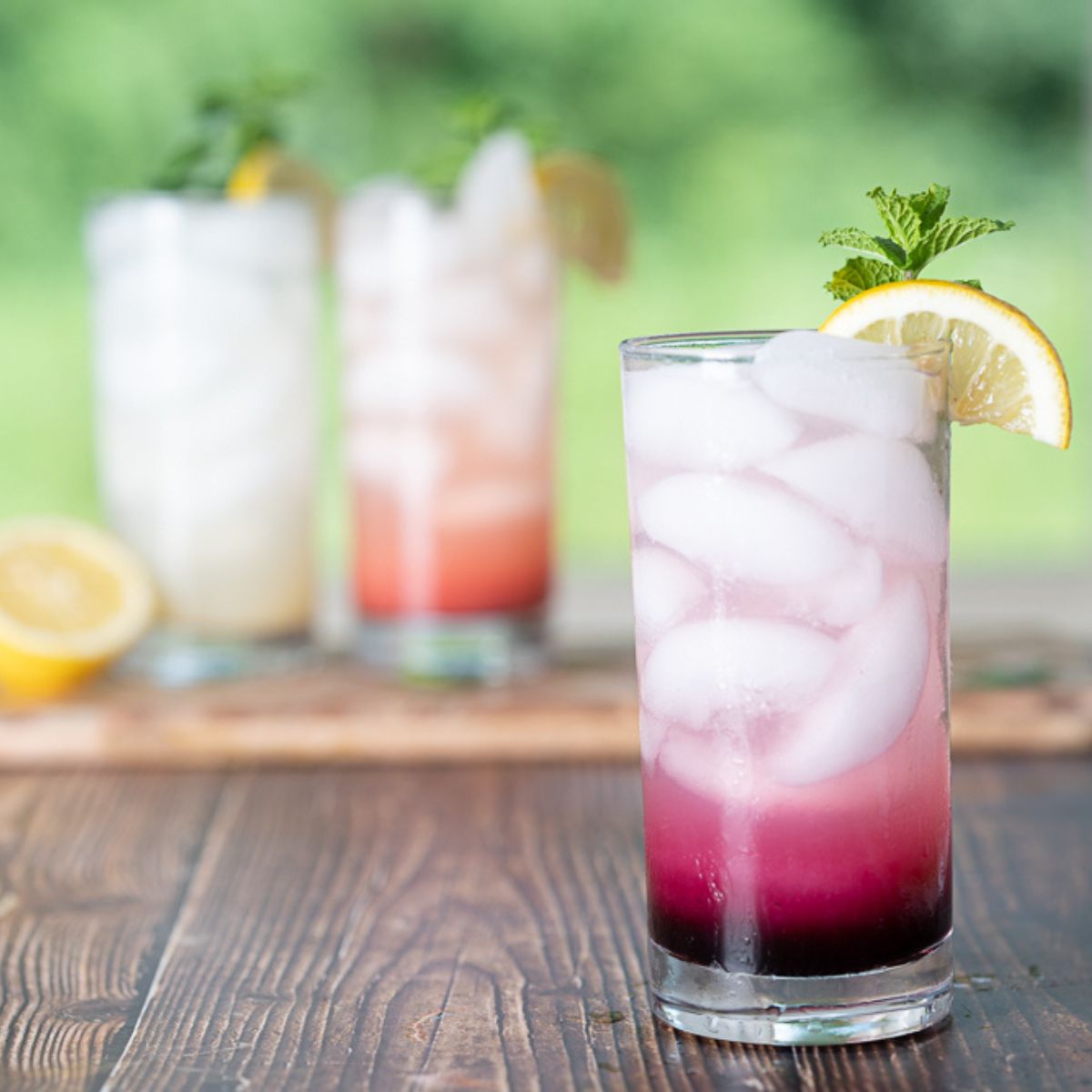 three mocktails with the blueberry mocktail in the front and a forest background behind the other two drinks